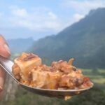 Spoonful of spicy mapo tofu held against the backdrop of Vang Vieng’s limestone mountains.